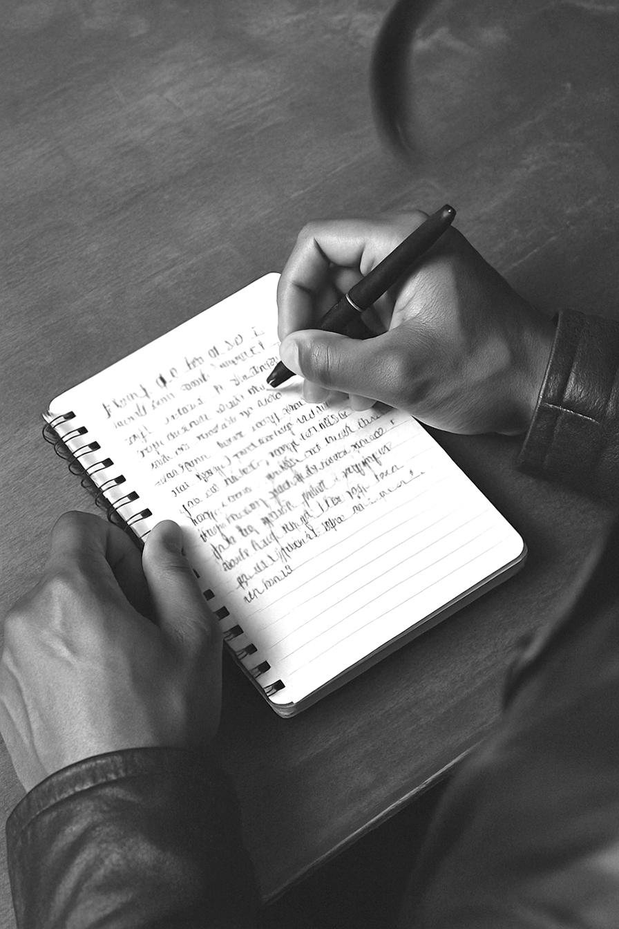 A hand writing longhand in a spiral notebook under a low lamp.